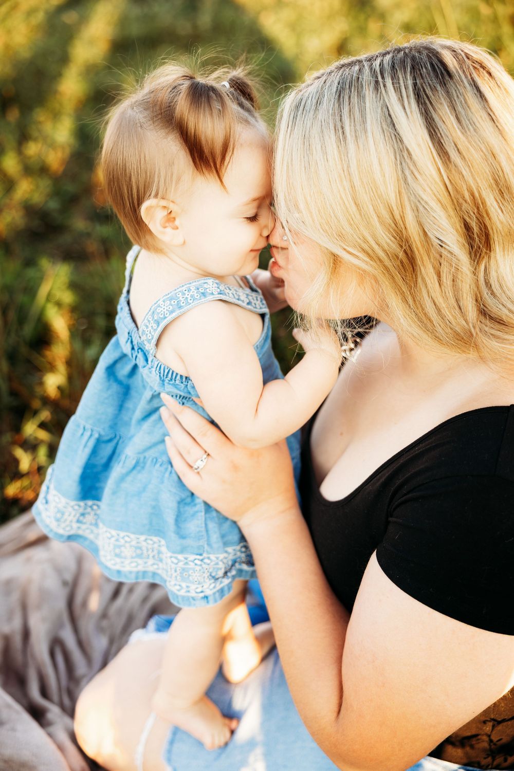 A blonde mother tenderly touches noses with her baby girl in a blue dress outdoors in warm sunlight.