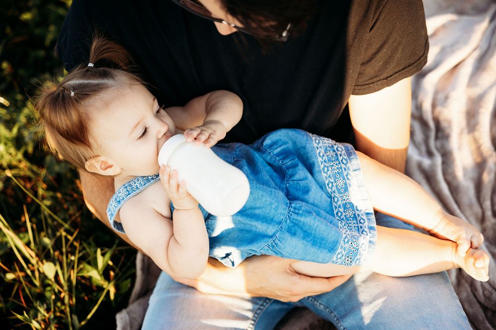 A mother cradling her baby girl in a blue dress while feeding her from a bottle outdoors in warm sunlight.