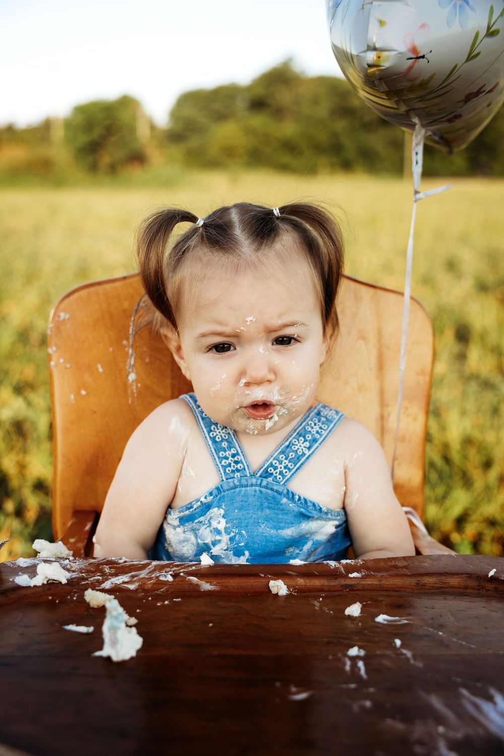 Messy baby girl in denim overalls eating smash cake in high chair outdoors with balloon on first birthday.