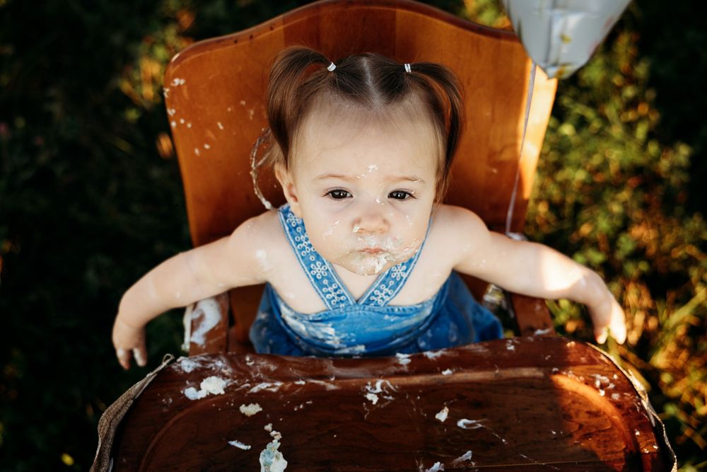Baby girl in blue overalls sitting in a wooden high chair covered in birthday cake frosting outdoors.
