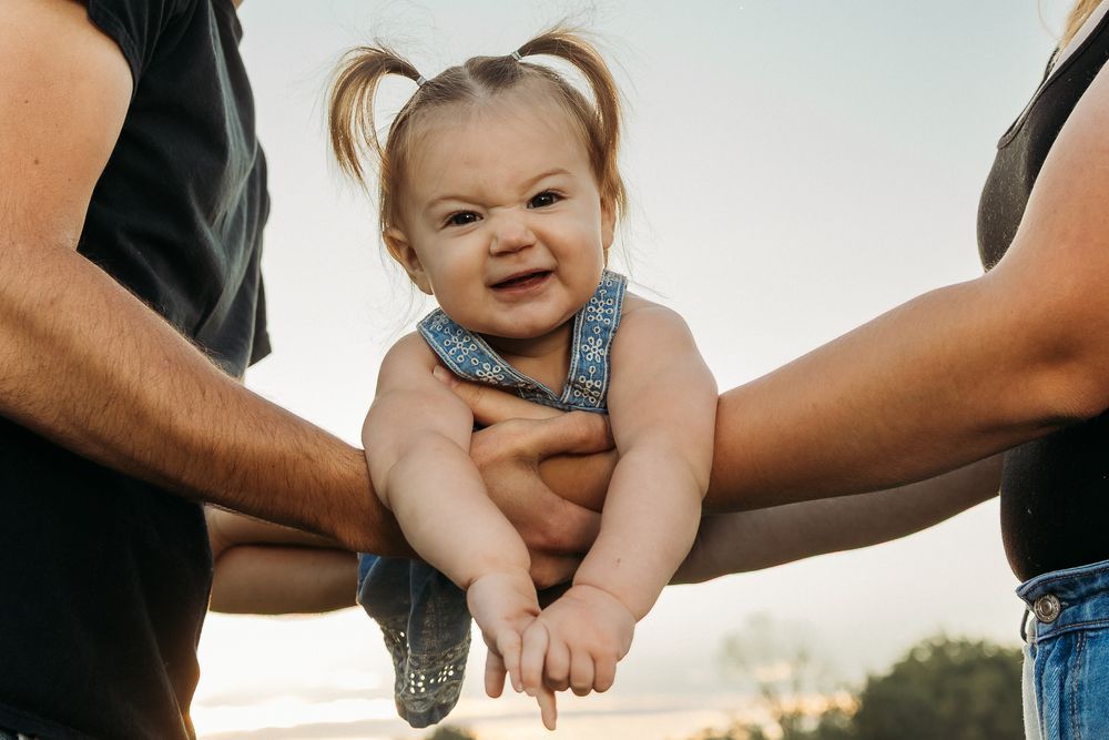 Two parents holding a smiling baby girl with pigtails in denim overalls outdoors at sunset.