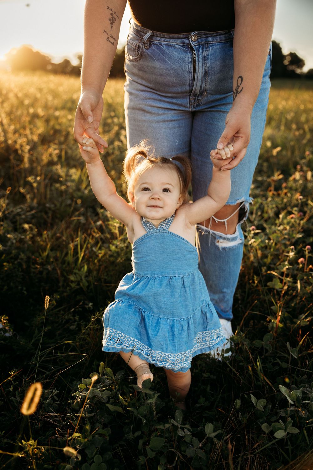 Toddler girl in blue dress holds mother's hands in sunlit field at golden hour.