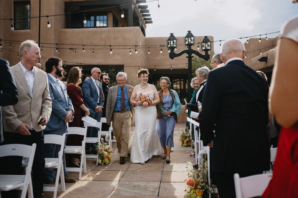 Bride walks down outdoor wedding aisle with escorts, guests seated in white chairs, string lights overhead.