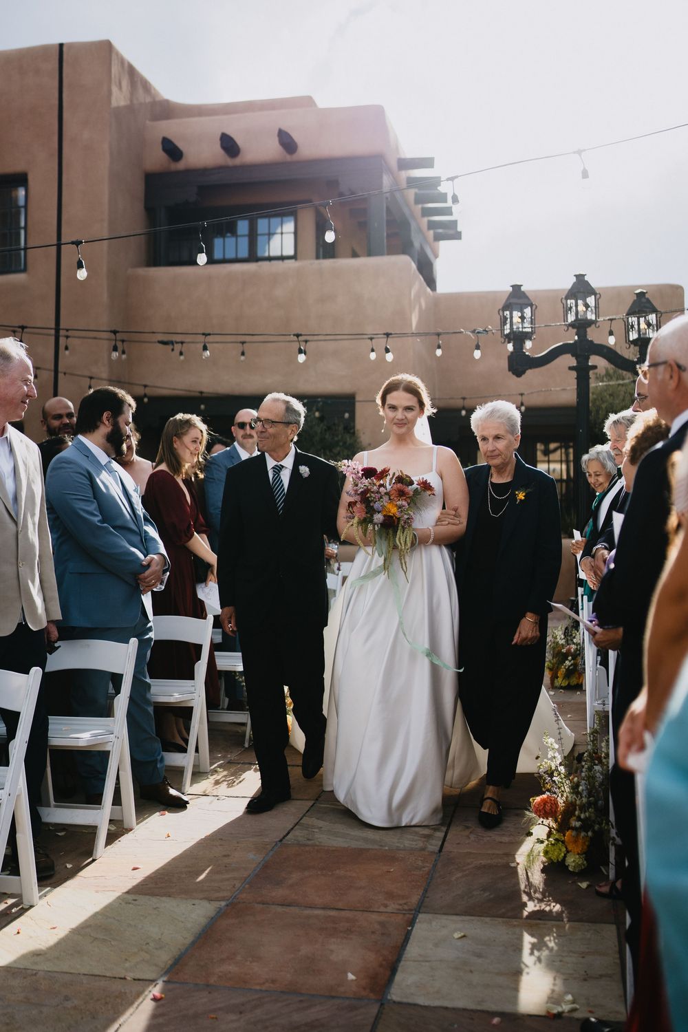 Bride walks down outdoor aisle with family, holding colorful bouquet, at a sunlit adobe-style wedding venue.
