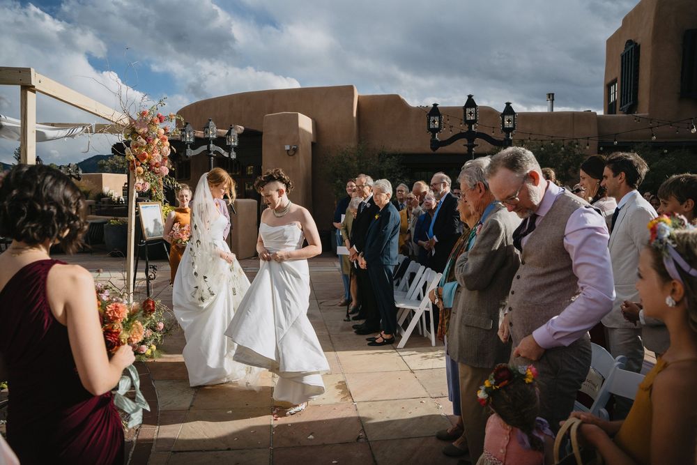 Bride walks down outdoor aisle in white gown at Southwest adobe venue wedding ceremony with guests seated.
