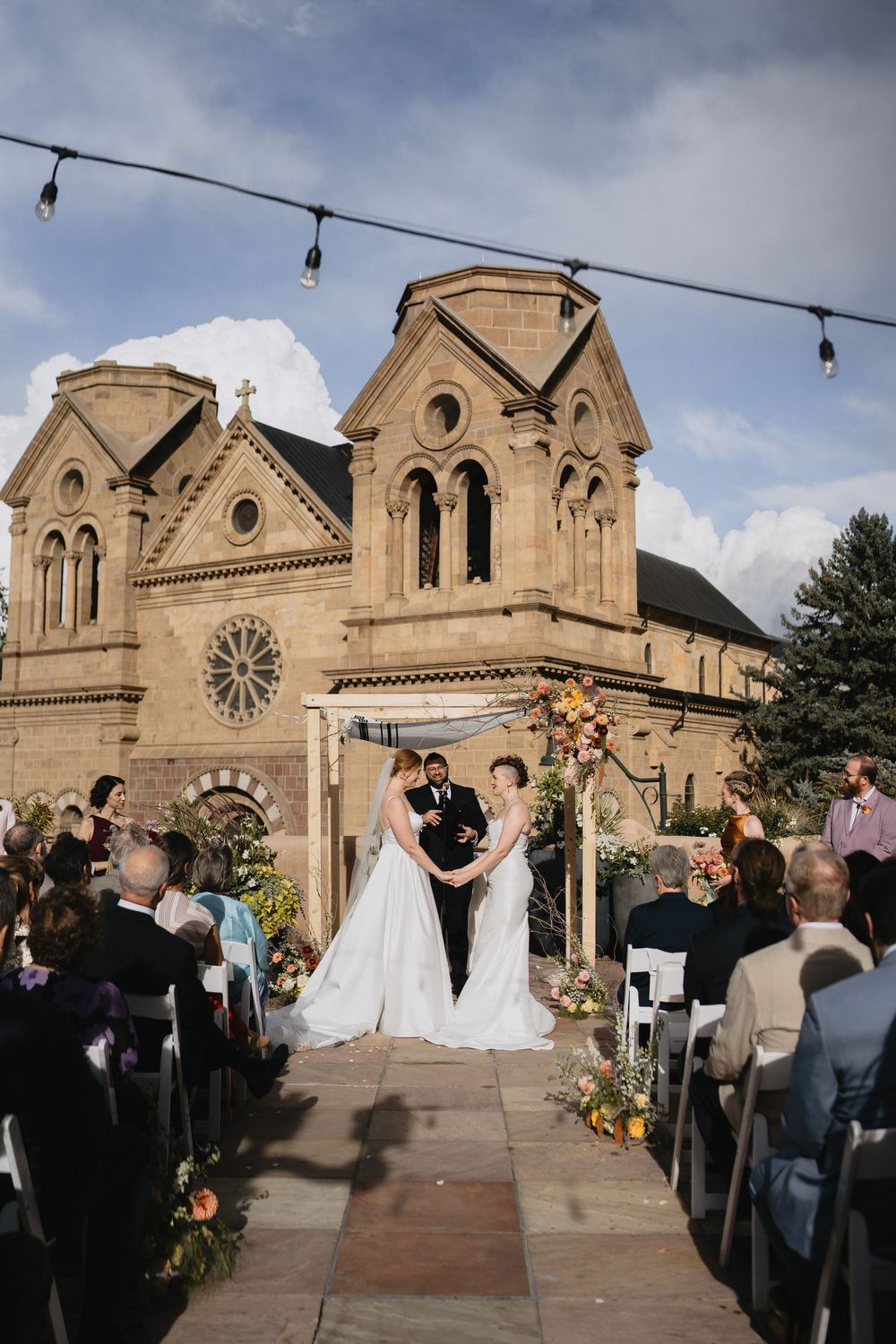 Two brides exchange vows outdoors before a stunning stone cathedral in Santa Fe, NM, with string lights overhead.