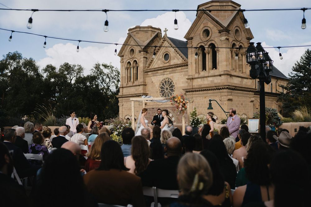 Outdoor wedding ceremony in front of historic stone cathedral with string lights and guests seated in Santa Fe, NM.