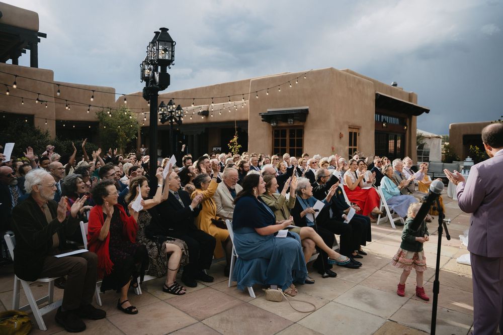 Large outdoor gathering of attendees raising hands at a Southwest-style adobe venue under cloudy skies.