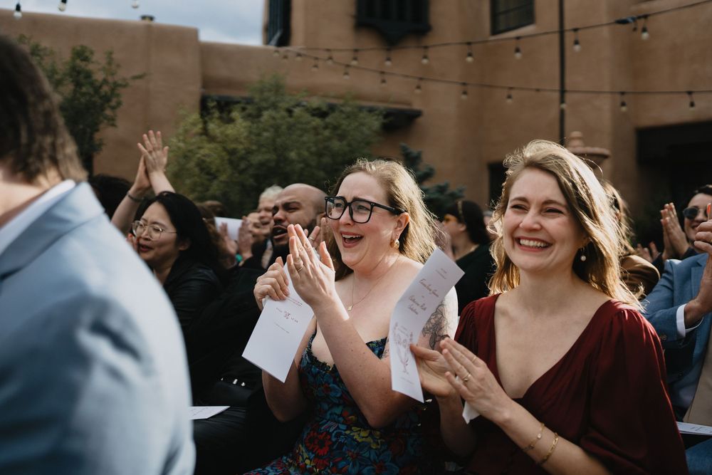 Guests clapping and smiling at an outdoor wedding ceremony, holding programs in a courtyard setting.