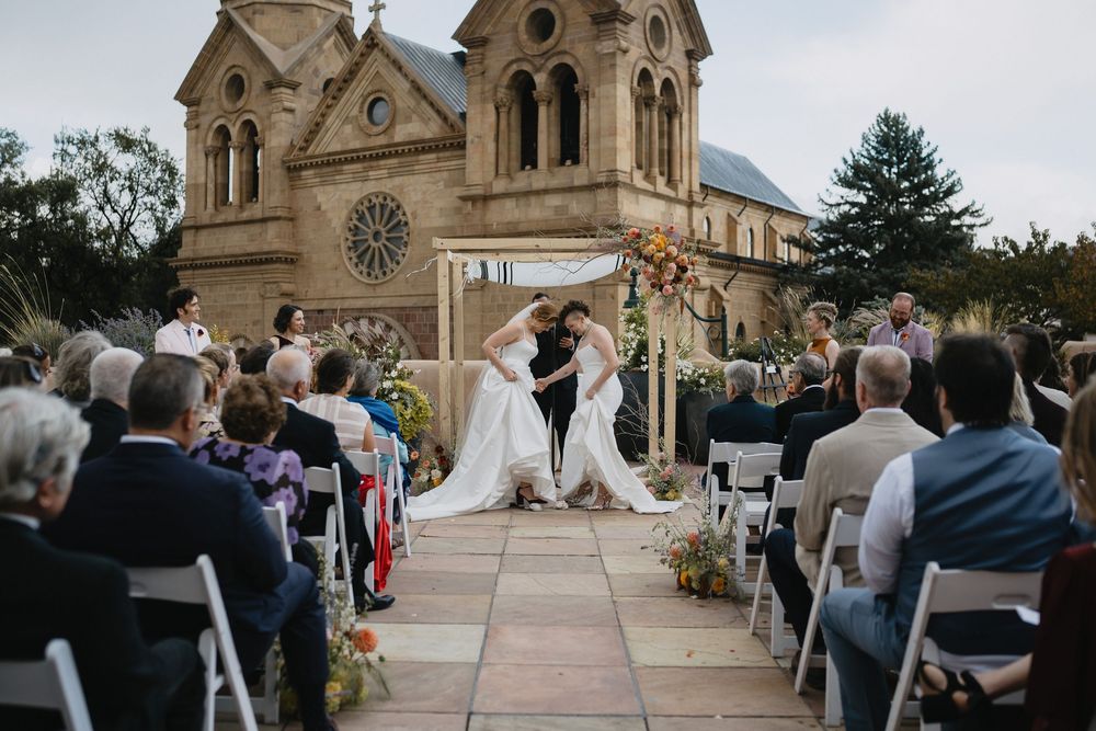 Two brides share a kiss at an outdoor wedding ceremony in front of a historic stone cathedral in Santa Fe.