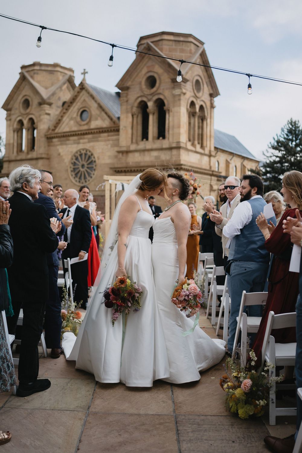 Two brides share a kiss at their outdoor wedding ceremony with a historic stone church backdrop and applauding guests.