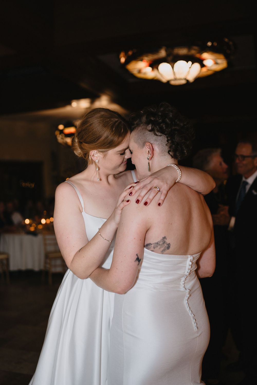 Two brides share an intimate first dance, embracing closely in elegant white wedding gowns at a dimly lit reception.