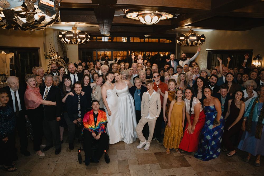 Large wedding group photo with bride and groom surrounded by smiling guests in elegant venue.