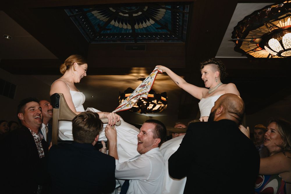 Bride and groom lifted by guests during lively wedding reception hora dance celebration indoors.