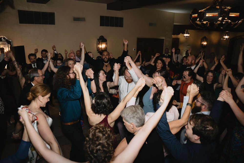 Crowd of wedding guests dancing with hands raised at a lively indoor reception venue.