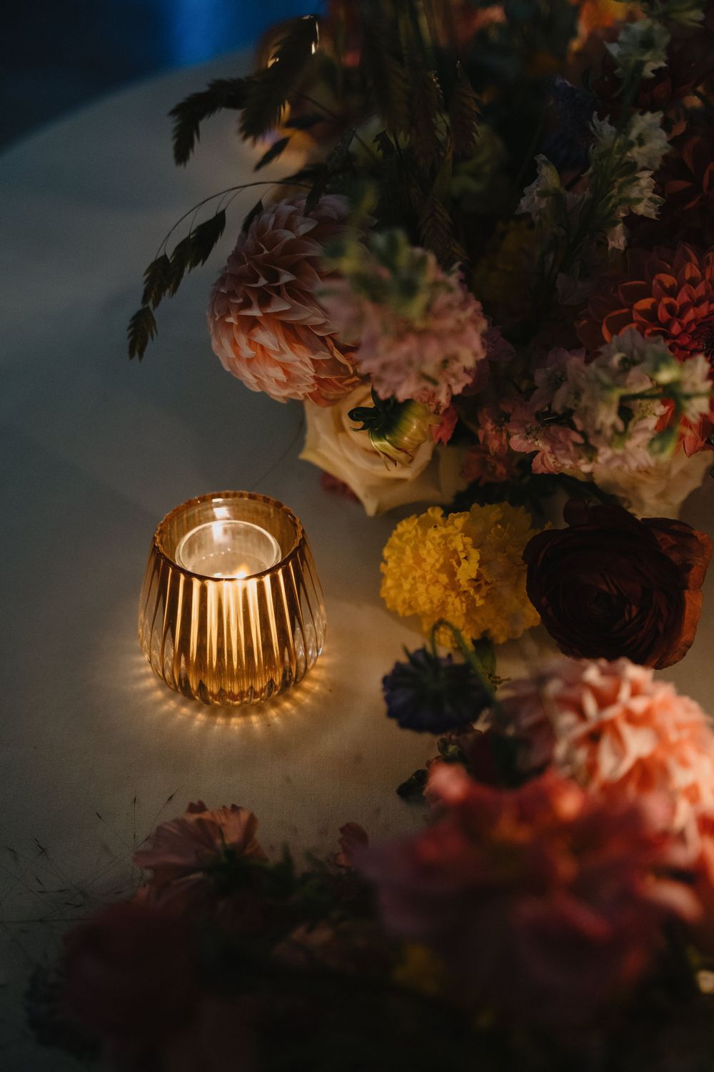 A glowing amber ribbed votive candle beside colorful floral arrangement with pink dahlias and marigolds in dim romantic light.