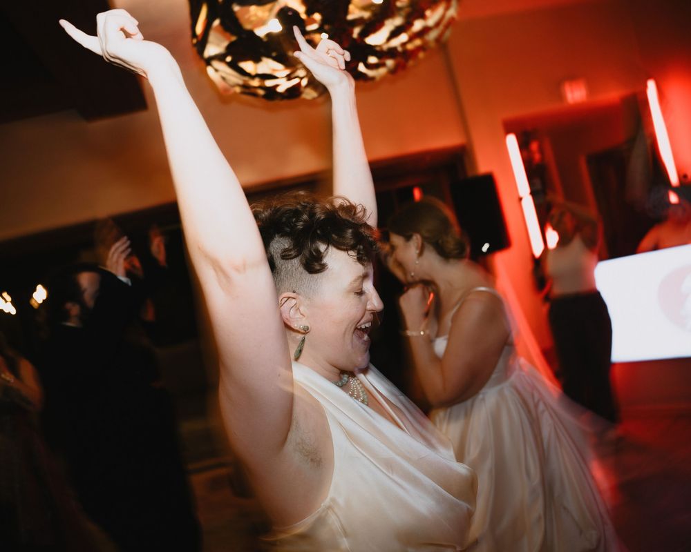 Joyful bride dancing with arms raised at wedding reception with red ambient lighting.