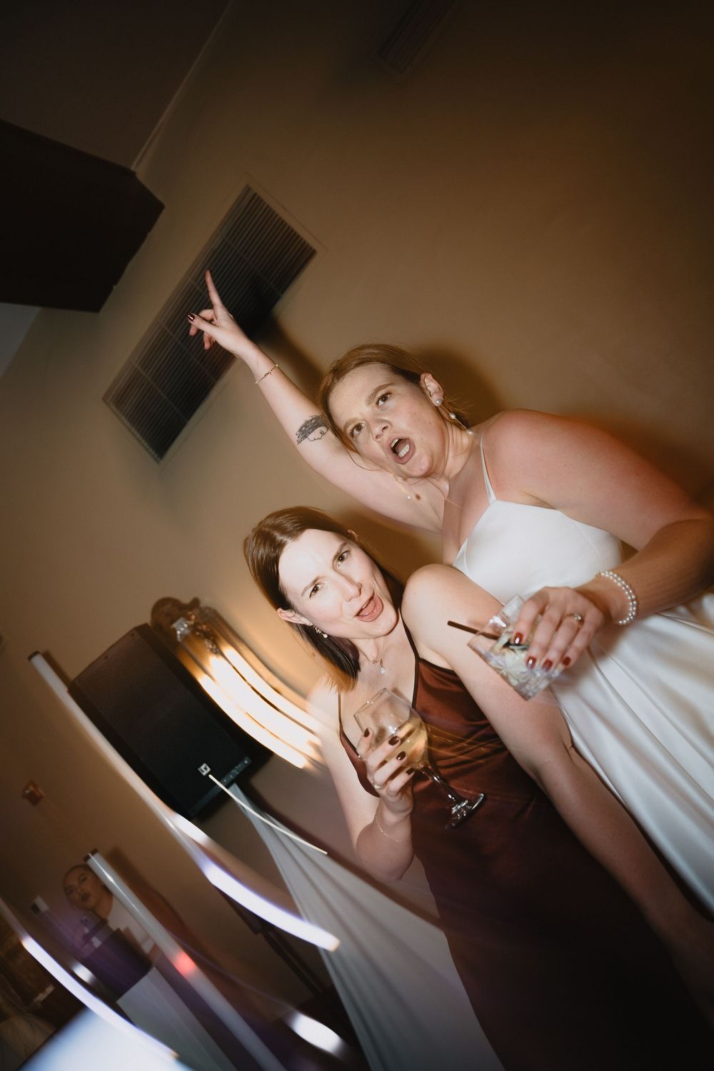 Two women in formal dresses celebrate with champagne glasses, one pointing upward, at what appears to be a wedding event.