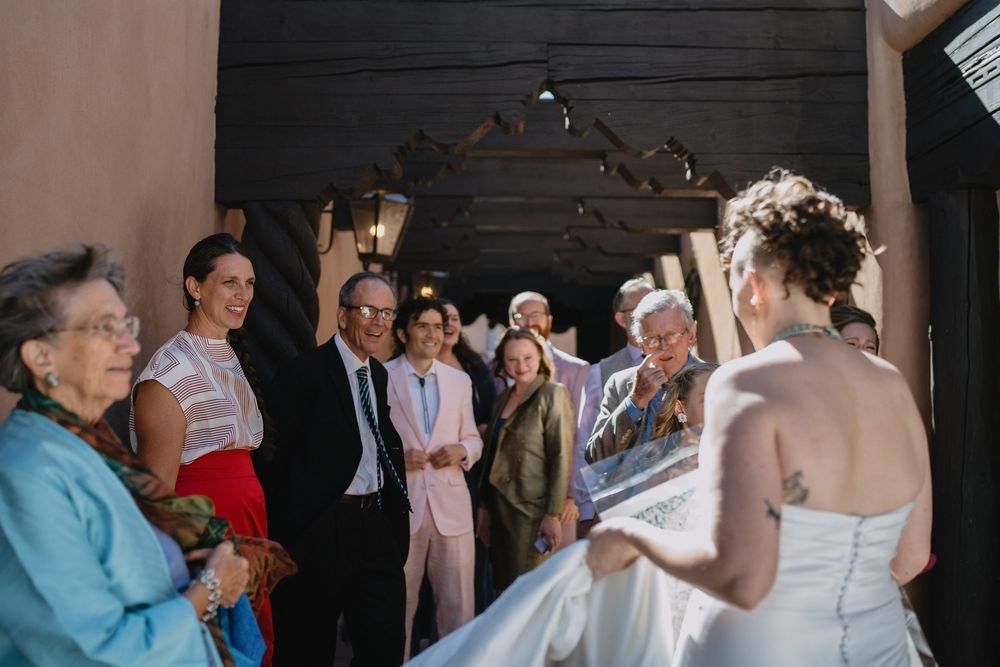 Bride in white gown shows dress to smiling wedding guests gathered at rustic venue entrance.