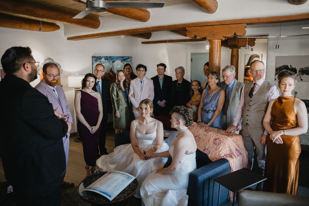 Two brides sit together during intimate wedding ceremony surrounded by small group of guests in rustic adobe room.
