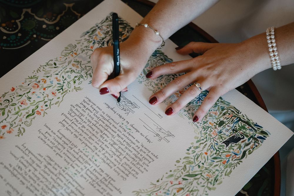 Bride signs ornate floral ketubah marriage contract with pen, wearing pearl bracelet and red nails.