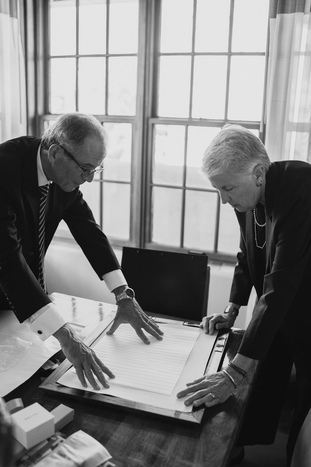 Two senior professionals in formal attire review documents together at a desk near large windows.