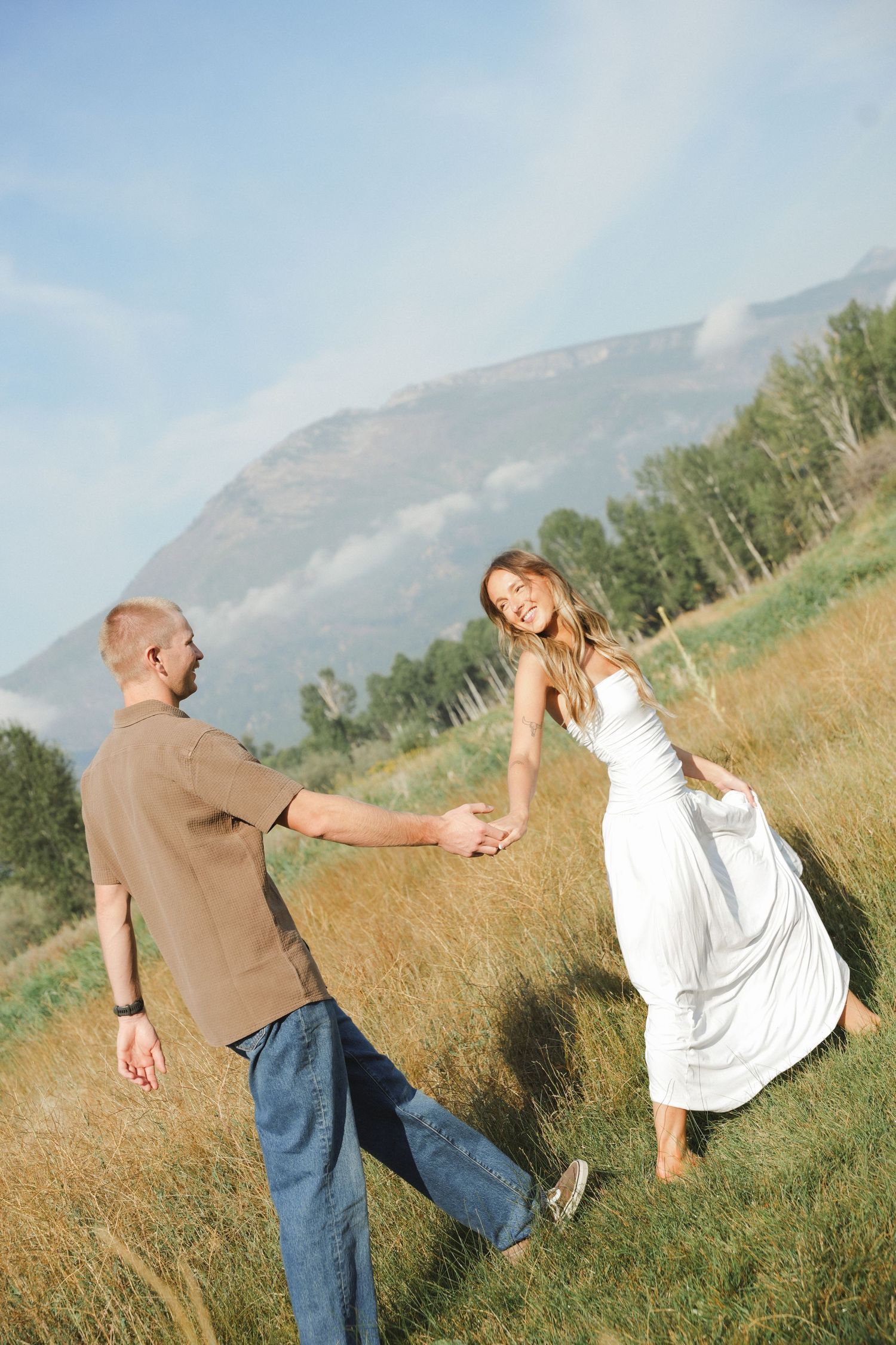 Couple sharing a romantic moment outdoors, man in gray cap gently touching woman's face near pine trees.