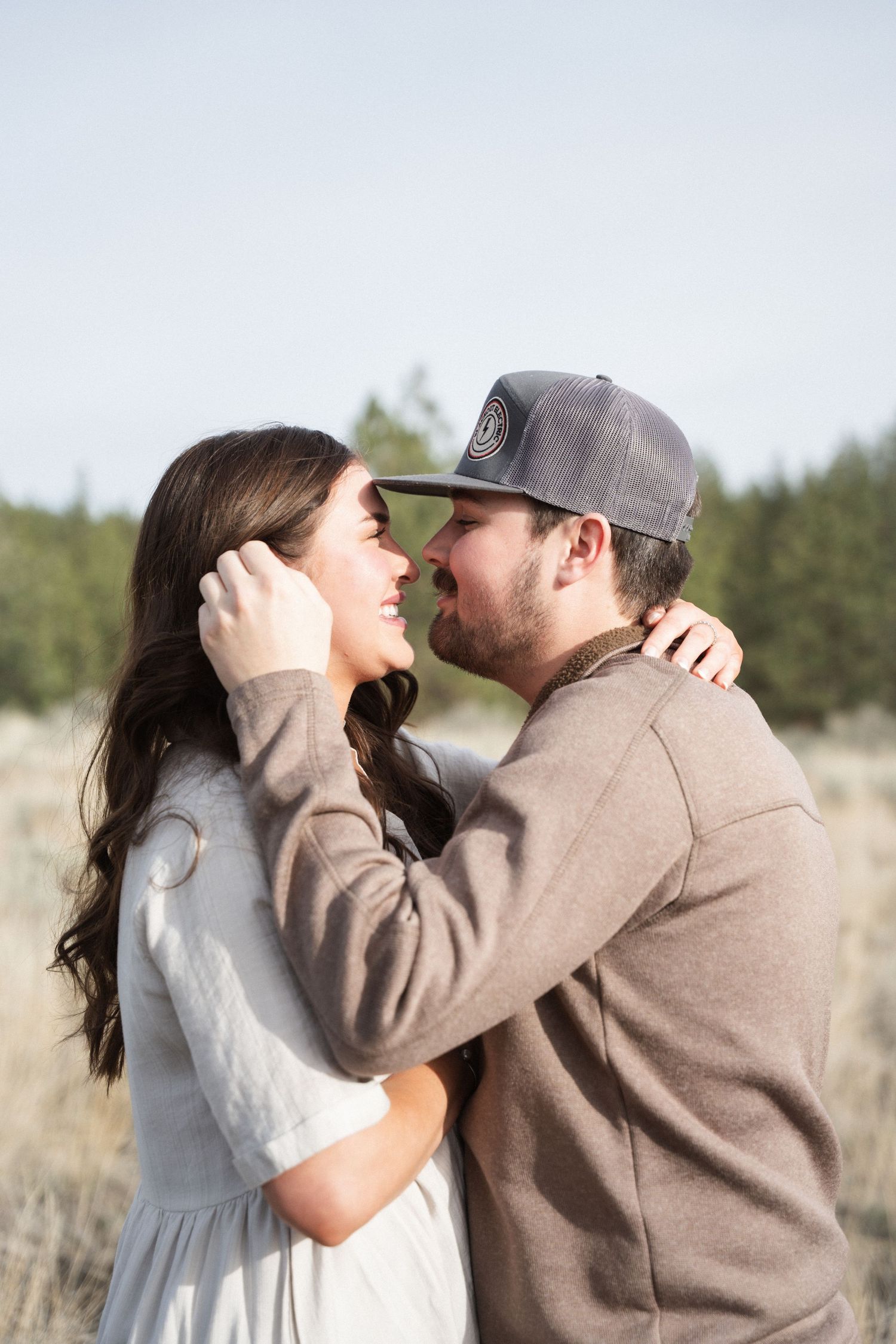 Couple sharing a romantic moment outdoors, man in gray cap gently touching woman's face near pine trees.