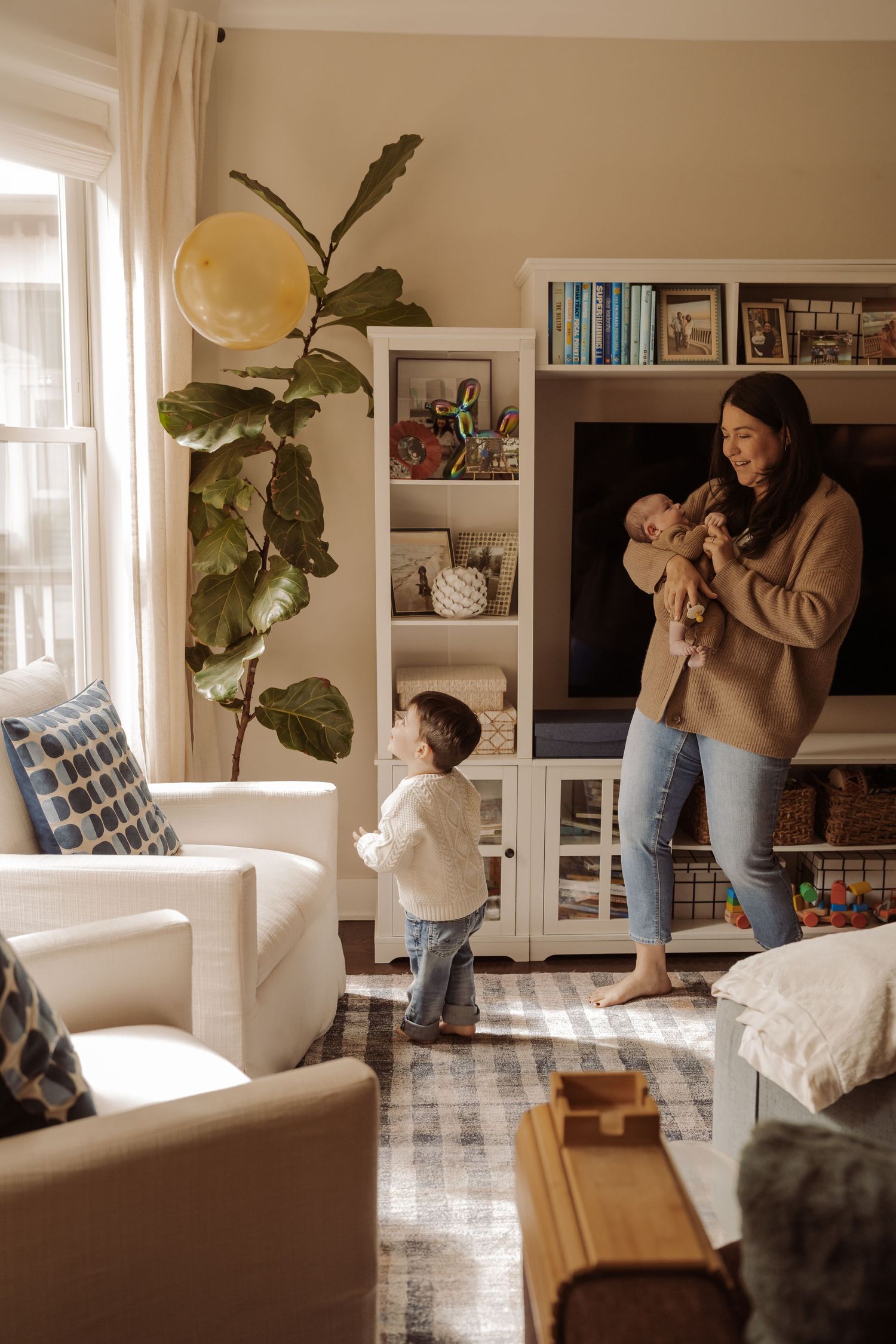 A mother holds her newborn while her toddler plays in a cozy living room with bookshelves and a fiddle leaf fig tree at a newborn session