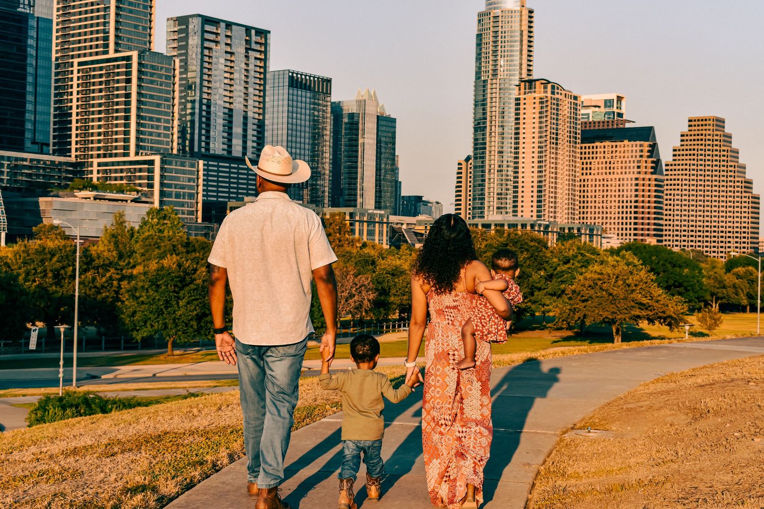 Family of four walks along a sunny park path with the Austin, Texas skyline visible in the background.