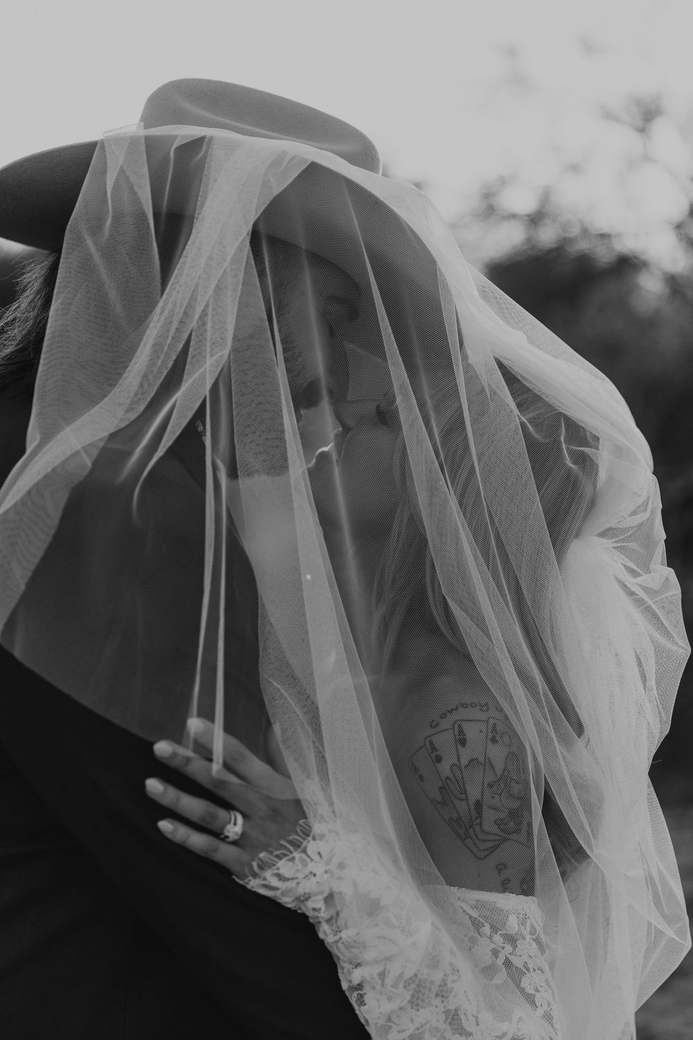 Black and white photo of a bride in lace dress with veil draped over her, revealing a tattoo and wedding ring.