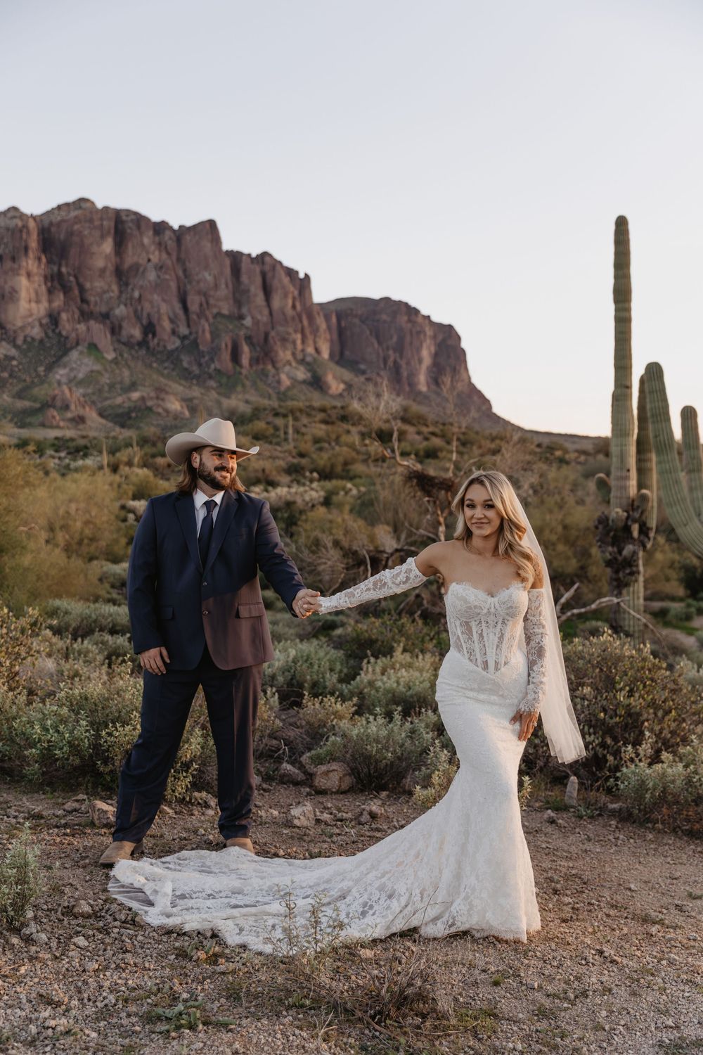 Bride and groom hold hands in Arizona desert with Superstition Mountains and saguaro cactus backdrop at sunset.