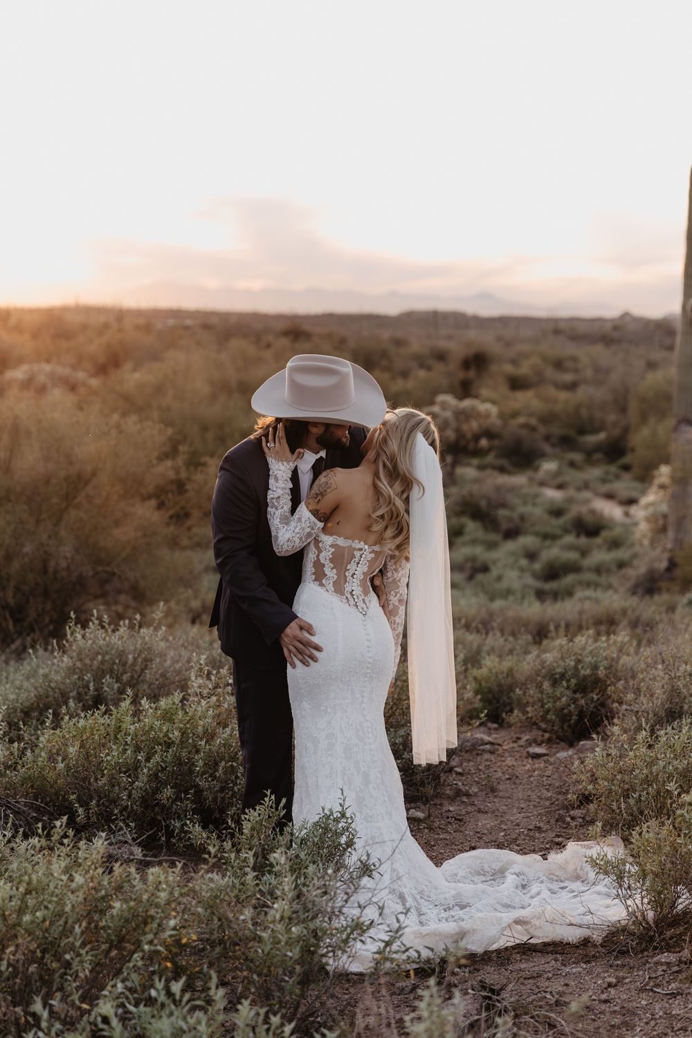 Bride and groom kissing at sunset in desert landscape, bride wearing lace gown with veil and groom in cowboy hat.