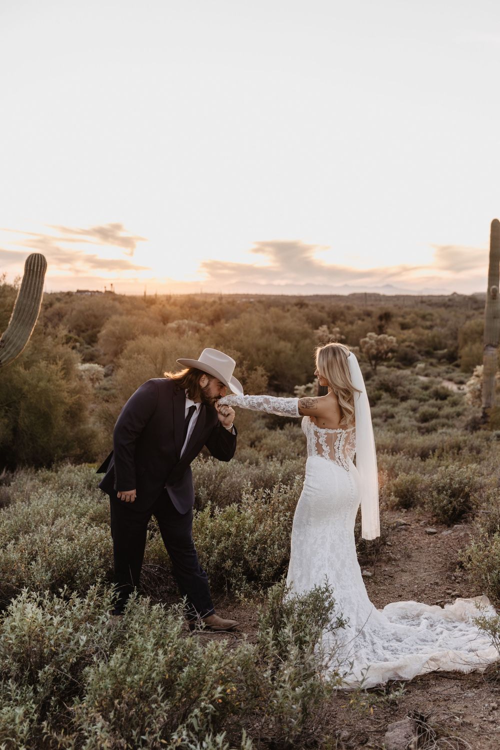 Groom in cowboy hat kissing bride's hand at sunset in Arizona desert with saguaro cacti backdrop.