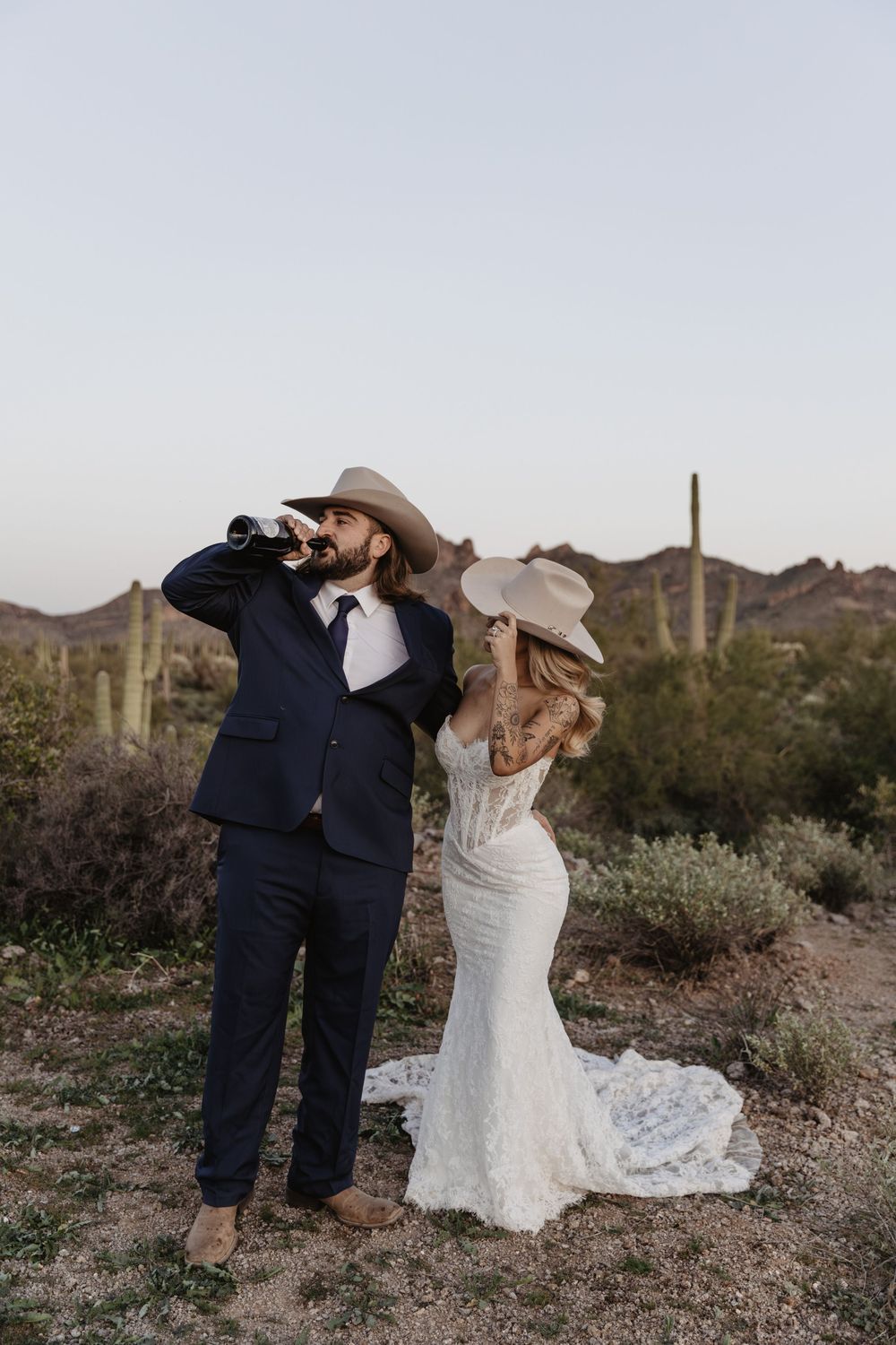 A bride and groom in cowboy hats celebrate in the Arizona desert, with saguaro cacti in the background.