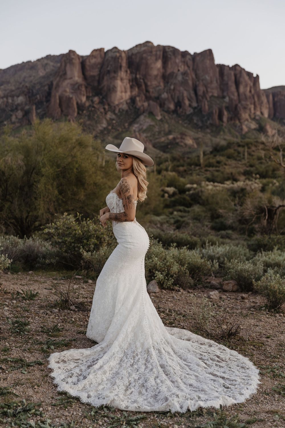 Bride in white lace mermaid gown and cowboy hat poses before rugged desert mountain landscape.