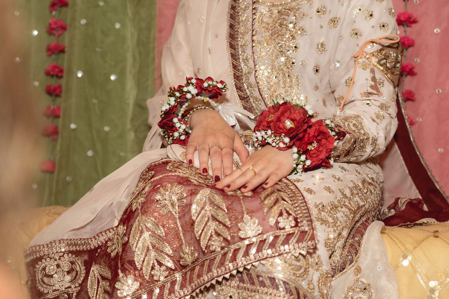 Pakistani bride in white and gold embroidered bridal outfit with red floral hand jewelry at wedding ceremony.