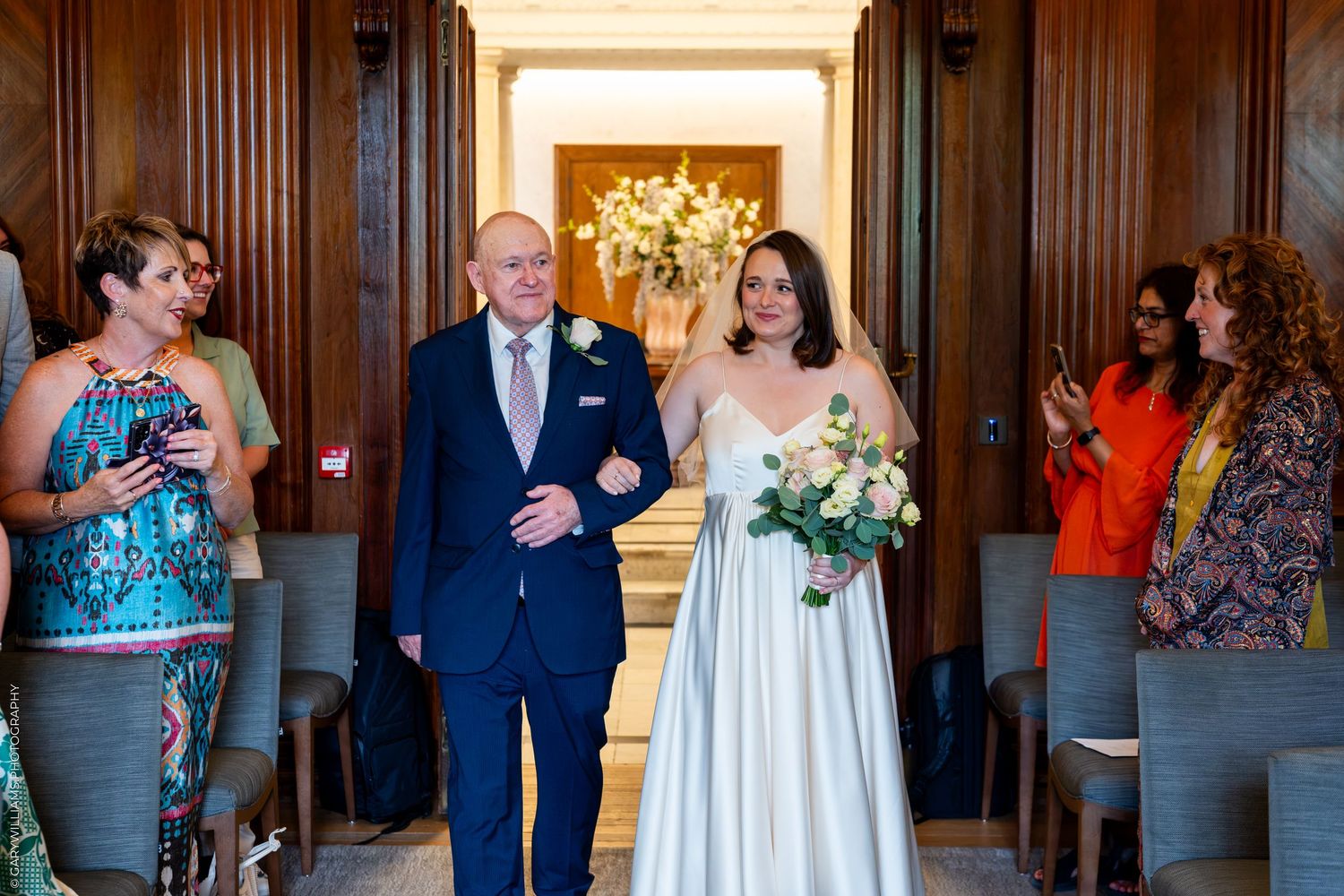 Bride in white gown walks down aisle with escort at elegant wood-paneled wedding venue, guests applauding.