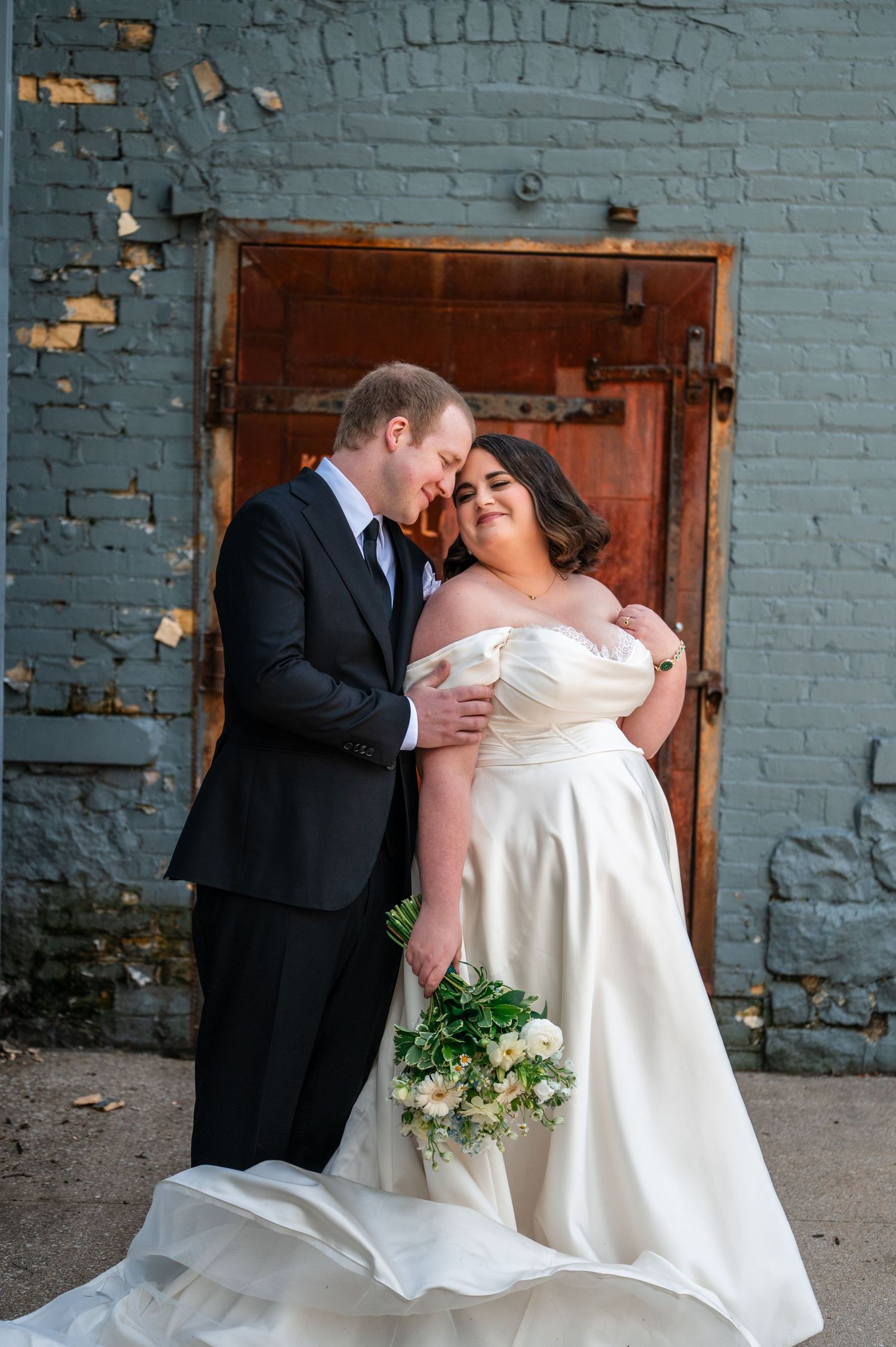 Bride in off-shoulder gown holding bouquet embraces groom in black suit before rustic brick wall.
