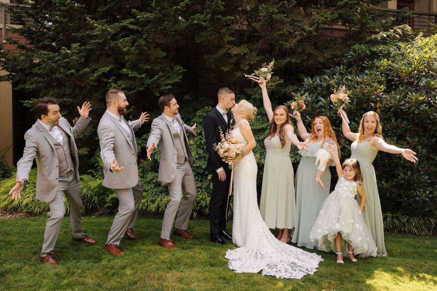 Wedding party celebrates outdoors, bridesmaids in sage green, groomsmen in gray suits, as bride and groom share a kiss.