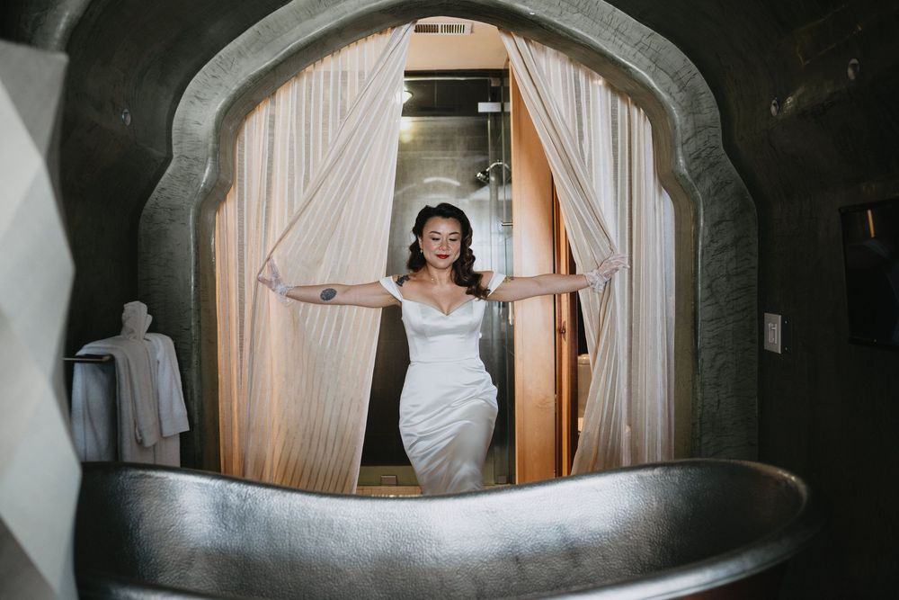 Smiling bride in white off-shoulder gown poses in an ornate arched doorway with curtains.