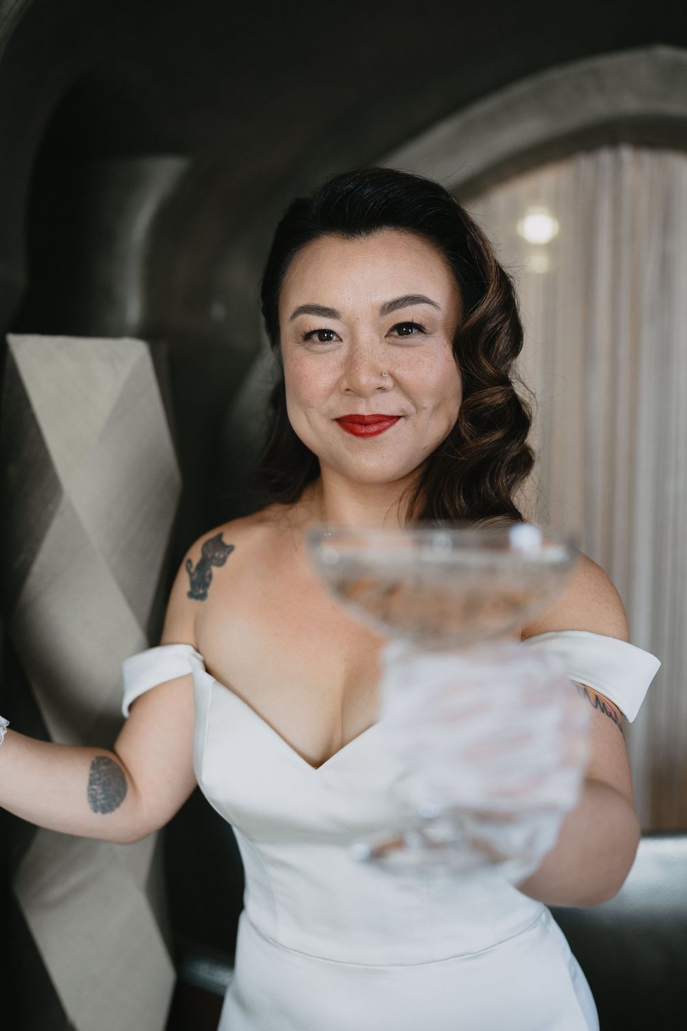 Tattooed bride in white off-shoulder gown holds a crystal glass and smiles at the camera.