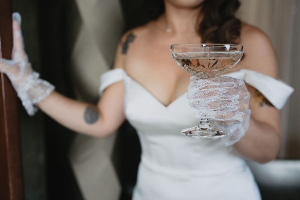 Close-up of tattooed bride in white gown holding a crystal coupe glass with lace gloves.
