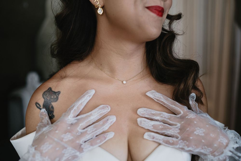 Close-up of bride wearing floral lace gloves with a cat tattoo on her shoulder in wedding dress.