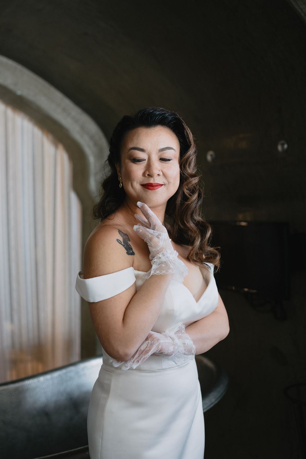 Elegant bride in white gown with lace gloves and red lips poses with eyes closed.