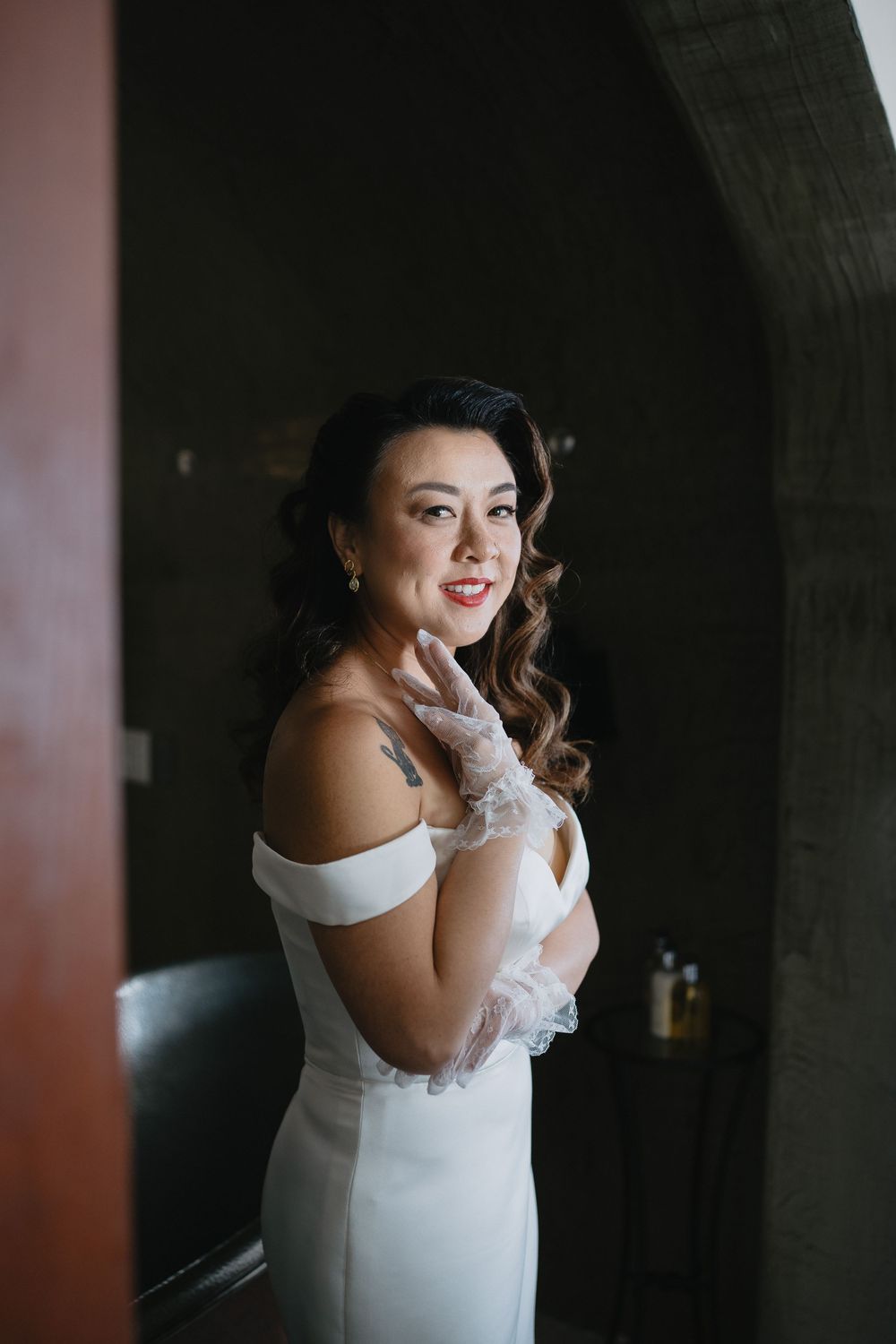 Radiant bride in off-shoulder white dress and lace gloves smiles in a dimly lit room.