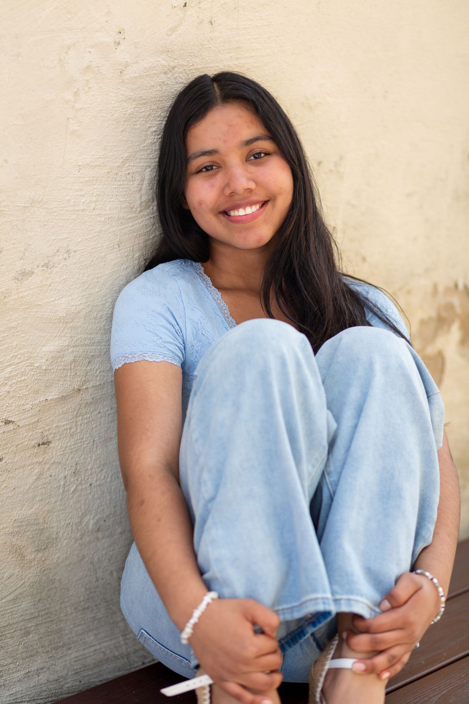 Smiling teenage girl in blue t-shirt and jeans sitting against a wall with knees up, wearing bracelets.