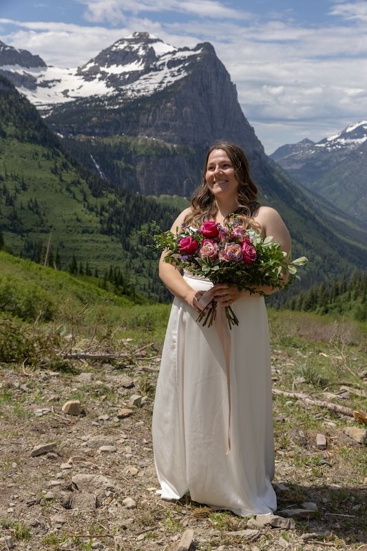 Smiling bride in white gown holds pink bouquet with snow-capped mountains and green valleys behind her.