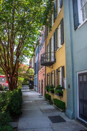 Colorful historic row houses with black shutters and balconies line a shaded sidewalk in Charleston, SC.