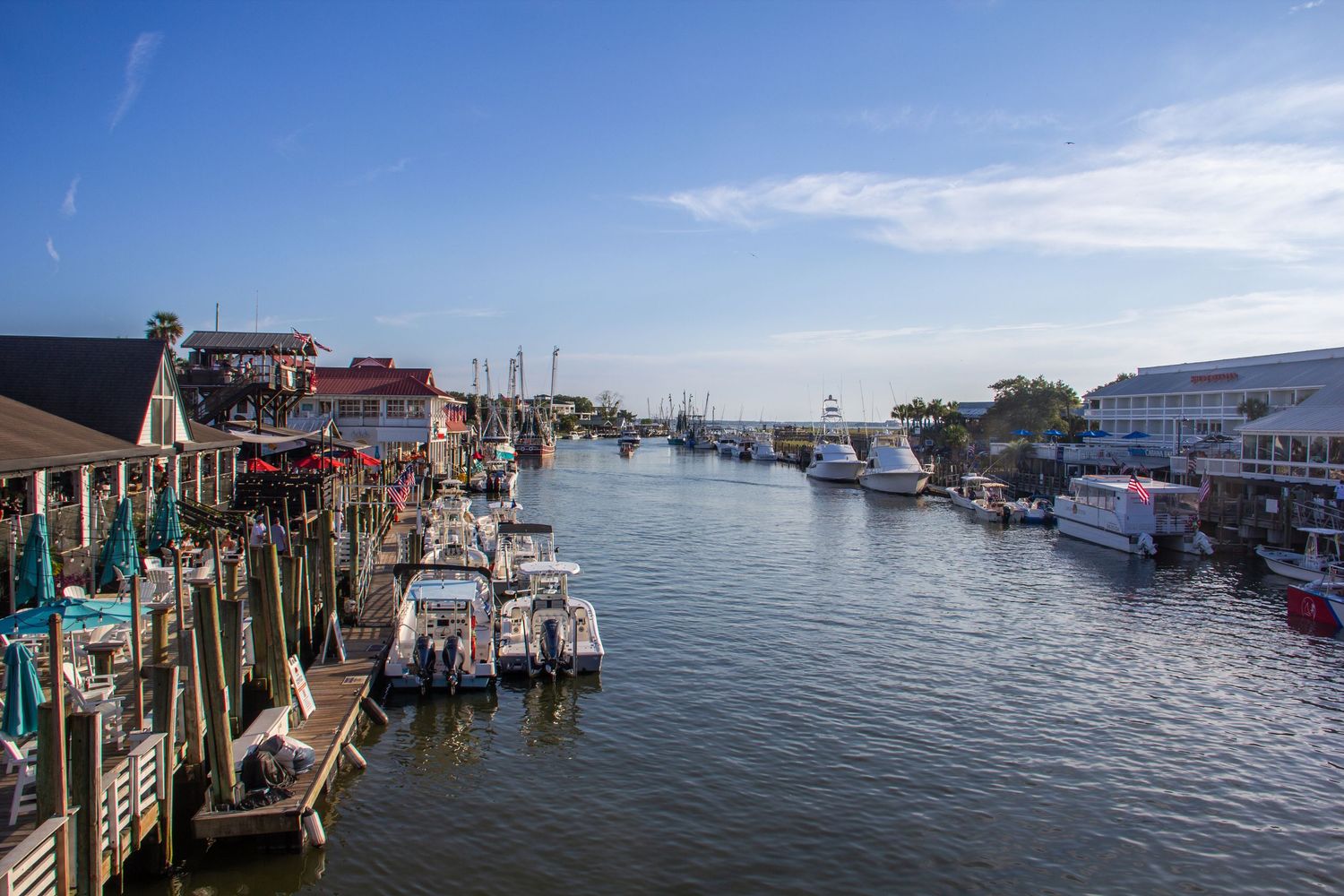 Scenic marina with boats docked along wooden piers flanking a calm waterway lined with waterfront shops under blue skies.